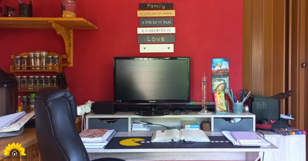Home desk beside a kitchen with an open Bible, planner, and notebooks arranged neatly beneath a wall sign about family and love.